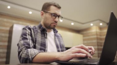 Beyaz sakallı genç adam mutfaktaki masada oturuyor ve uzaktan çalışmak için dizüstü bilgisayar kullanıyor. Ellere odaklan. İnternette çalışıyorum. Yavaş çekim