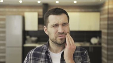 Mutfakta otururken diş ağrısı çeken mutsuz, genç sakallı bir adam. İnsanlar, sağlık hizmetleri, dişçilik ve sorun konsepti. Yavaş çekim