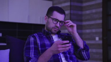 Evde oturma odasında otururken cep telefonu ekranına bakan gözlüklü sakallı sakin genç adam. Yaşam tarzı konsepti. Yavaş çekim