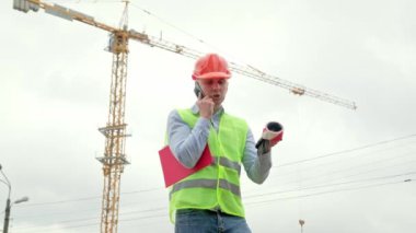 Kolunun altında pano olan ciddi bir ustabaşı kule vincinin önünde akıllı telefondan konuşuyor. Yavaş çekim