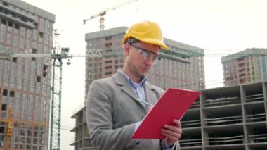 Kule vinçleri ve bitmemiş çok katlı evlerin yanında dururken site yöneticisinin belden yukarı portresi panoya yazılmış. Yavaş çekim