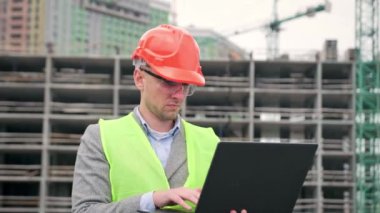Odaklanmış site yöneticisi tamamlanmamış çok katlı binaların yanında dururken taşınabilir bilgisayara veri giriyor. Yavaş çekim