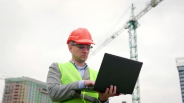 Ciddi inşaat denetçisi, kule vinci ve tamamlanmamış çok katlı evlerin yanında dururken verileri dizüstü bilgisayara giriyor. Yavaş çekim