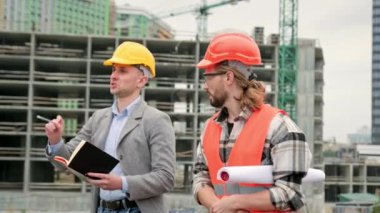 İnşaat müdürü, not defterine danışırken ustabaşına bir şey işaret ediyor ve teftiş sırasında konuşuyor. Yavaş çekim