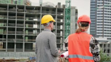 Site yöneticisi bitmemiş bir eve doğru yürürken ustabaşıyla konuşuyor. Yavaş çekim