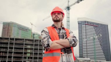 Ciddi bir ustabaşı, eli kolu bağlanmış, bitmemiş bir konutun yakınında duruyor ve uzaklığa bakıyor. Yavaş çekim