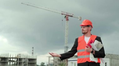 Ofis kostümü içinde gülümseyen bir müteahhit ve elinde pano olan koruyucu turuncu bir miğfer, planlar ve gösterim alanı. Yavaş çekim