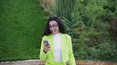 Yeşil ofis kıyafetleri içinde, elinde akıllı telefon olan ve kazanmaktan zevk alan, parkta yürüyen bir kadın. Yavaş çekim