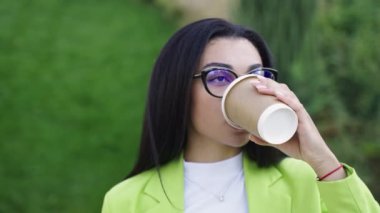 Yeşil ofis elbiseli bir kadın, parkta duruyor ve kahve içiyor. Yavaş çekim videosu