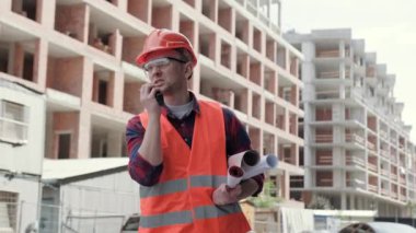 Kolunun altında sarılmış mimari çizimleri olan bir inşaatçı bitmemiş çok katlı binaların arasında dikilirken telsizle konuşuyor. Yavaş çekim