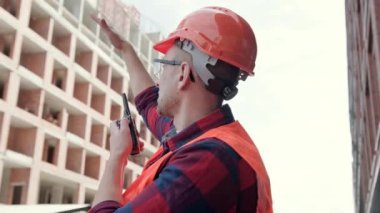 İnşaat müfettişi, iki yönlü telsiz üzerinden el kol hareketi yapıp iletişim kurarken yarım kalmış çok katlı evin önünde duruyor. Yavaş çekim
