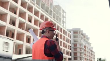 İnşaat müdürünün, bitmemiş çok katlı evlerin önünde dikilirken telsizle konuşan çizimlerinin arkası. Yavaş çekim