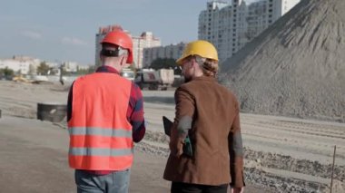 Foreman koruyucu miğfer ve kıyafetlerle şantiyede yürüyor ve müşteriyle konuşuyor. Yavaş çekim