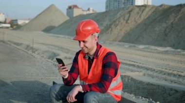 Koruyucu giysiler ve miğfer giyen inşaatçı inşaatta oturuyor, akıllı telefon tutuyor ve sohbet ediyor. Yavaş çekim