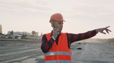 Koruyucu giysili ve kasklı bir adam telsiz tutuyor ve inşaatçılarla konuşuyor, talimatlar veriyor. Yavaş çekim