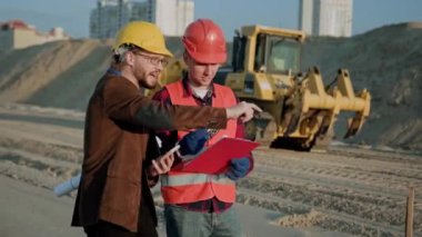 Koruyucu giysiler ve kask giyen ustabaşı müşteri belgelerini kontrol ediyor, kazıcı yakınlardan geçiyor. Yavaş çekim