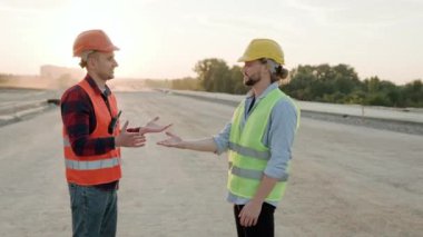 Koruyucu miğferler ve yelek giymiş inşaatçılar dışarıda dikilip mekanı inceliyor ve tartışıyorlar. Yavaş çekim videosu