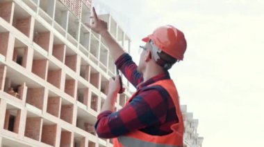 Koruyucu giysiler ve miğfer giymiş bina kenarında duran, elinde telsiz olan ve işçilerle konuşan bir inşaat işçisi. Yavaş çekim