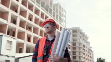 Koruyucu giysili ve miğferli bir inşaatçı binanın yanında duruyor. Elinde planlar, kontrol işleri var. Yavaş çekim