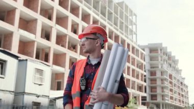 Koruyucu giysili ve miğferli bir bina inşaatçısı binanın yanında duruyor, elinde planlar var, tamam işareti gösteriyor. Yavaş çekim