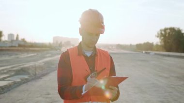 Koruyucu giysiler ve miğfer takmış bir müşteri elinde dosya tutuyor, belgeleri kontrol ediyor, notlar alıyor. Yavaş çekim