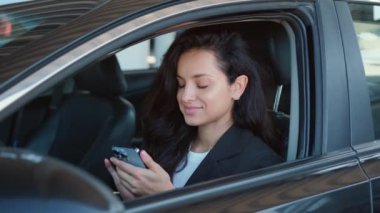 Açık camlı arabada oturan, rahatlayan, akıllı telefonda yazan bir kadın portresi. Yavaş çekim videosu