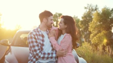 Gülümseyen adam ve kadın birbirlerine bakıyor, bir arabanın kaputuna yaslanırken sarılıyorlar. Ulaşım, seyahat, ilişki, insan konsepti. Yavaş çekim