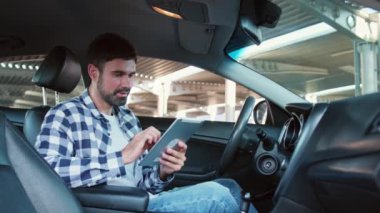 Genç adam arabada otururken dijital tablet üzerinde yazıyor. Ulaşım, teknoloji, yaşam tarzı konsepti. Yavaş çekim