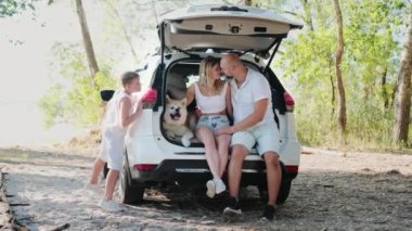 Açık bagajda Akita Inu ile oturan gülümseyen bir çift, arabanın arkasına saklanan küçük bir çocuk. Aile, yaz tatili konsepti. Yavaş çekim