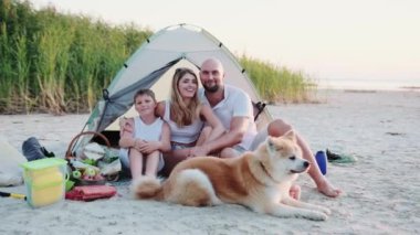 Gülümseyen genç aile ve köpek çadırda otururken sahilde piknik yapıyorlar. Aile, yaz tatili konsepti. Yavaş çekim