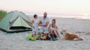 Gülümseyen ebeveynler ve küçük çocuk piknik sırasında sahilde otururken içkinin tadını çıkarıyorlar. Ağır çekim. Aile, eğlence ve insan konsepti