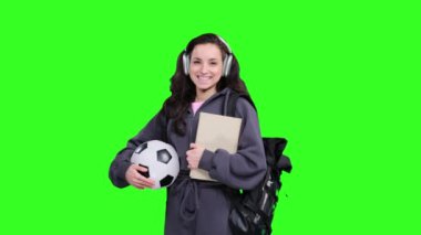 Sırt çantalı gülümseyen genç bir öğrenci futbol topu ve kitap ile poz verirken yeşil izole edilmiş yerde kulaklık kullanıyor.
