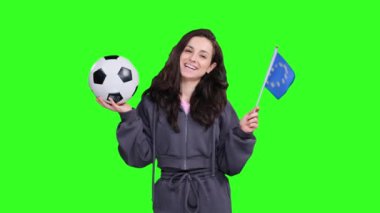Gülümseyen genç bayan hayran Avrupa Birliği bayrağının yanında poz veriyor ve sallanırken yeşil izole edilmiş bir yerde futbol topu tutuyor.