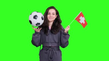 Yeşil izole edilmiş bir yerde elinde futbol topu tutarken İsviçre bayraklarıyla poz veren ve dalgalanan mutlu genç bayan hayranlar.