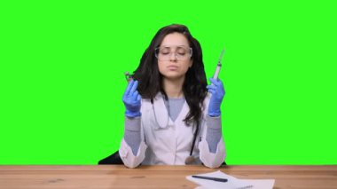 Koruyucu gözlüklü kadın doktor elinde aşı ve şırınga tutarken yeşil izole arka planda oturuyor. Sağlık hizmeti, tıp konsepti