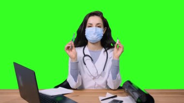 Korunaklı yüz maskesi takan kadın doktor, aşı ve şırıngayı nallarken aynı zamanda yeşil izole arka planda ofiste oturuyor. Sağlık hizmeti, tıp konsepti