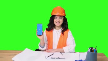 Koruyucu başlık takmış gülümseyen genç bayan mühendis telefon ekranı ve yeşil izole arka planda iyi bir hareket gösteriyor. Endüstri, mühendislik kavramı