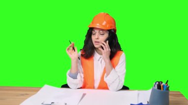 Korumalı miğferli genç bayan mühendis, yeşil izole arka plan notları alırken akıllı telefondan konuşuyor. Endüstri, mühendislik kavramı