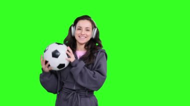 Gülen Kafkasyalı sporcu kadın futbol topunu tutarken kusuyor, kulaklıkla yeşil izole alanda müzik dinliyor.