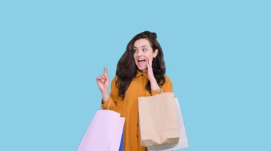 Alışveriş torbaları tutan mutlu genç bir kadın mavi izole arka plandaki boşluğu parmağıyla işaret ediyor. Kara Cuma, satış konsepti