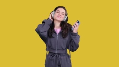 Güzel genç bir kadın akıllı telefon tutarken dans ediyor, kablosuz kulaklık takıyor, sarı izole edilmiş müzik dinliyor.