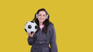 Sarı izole edilmiş bir yerde elinde futbol topu olan mutlu, sportif bir kadın müzik dinliyor.