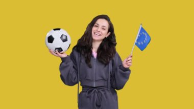 Gülümseyen genç bir kadın taraftar, sarı izole edilmiş bir futbol topunu tutarken Avrupa Birliği bayraklarıyla poz veriyor ve sallıyor.