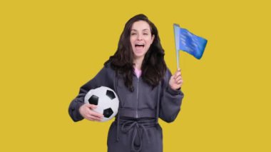 Bir futbol topu tutarken Avrupa Birliği bayraklarıyla poz veren genç ve mutlu bir bayan taraftar sarı izole edilmiş takımda takım zaferini kutluyor.