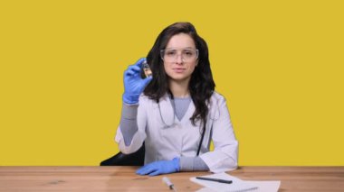 Koruyucu gözlüklü, elinde aşı şişesi olan üniformalı kadın doktor sarı izole edilmiş arka planda oturuyor. Sağlık hizmeti, tıp konsepti
