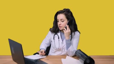 Üniformalı kadın doktor akıllı telefonla konuşurken masada oturuyor ve sarı izole edilmiş arka planda dizüstü bilgisayar kullanıyor. Sağlık hizmeti, tıp konsepti
