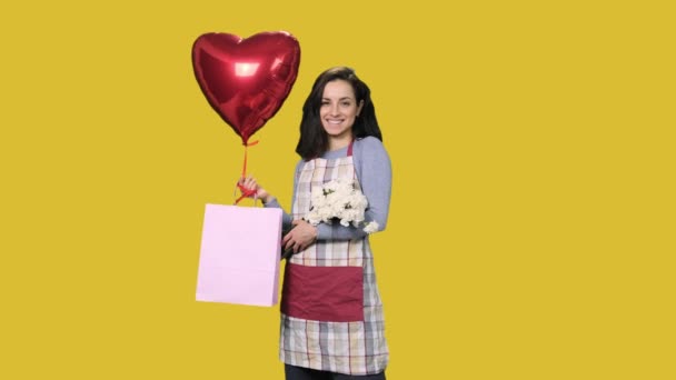 白いカーネーション 現在のバッグ 赤いハート型の気球を黄色の隔離された背景に保持する笑顔の女性の花屋 中小企業コンセプト — ストック動画