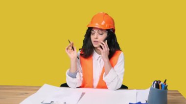 Korumalı miğferli genç bayan mühendis sarı izole arka plan notları alırken akıllı telefondan konuşuyor. Endüstri, mühendislik kavramı