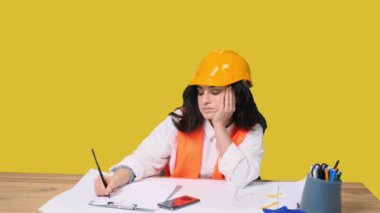 Güvenlik kaskı takmış uykulu kadın mimar masada oturuyor ve sarı izole arkaplanı çizmeyi bitiriyor. Endüstri, mühendislik kavramı
