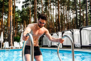 Açık havuzda poz verirken güneş gözlüğü takan kaslı adam. Arka planda ağaçlar var. Yaz zamanı, tatiller, yaşam tarzı konsepti